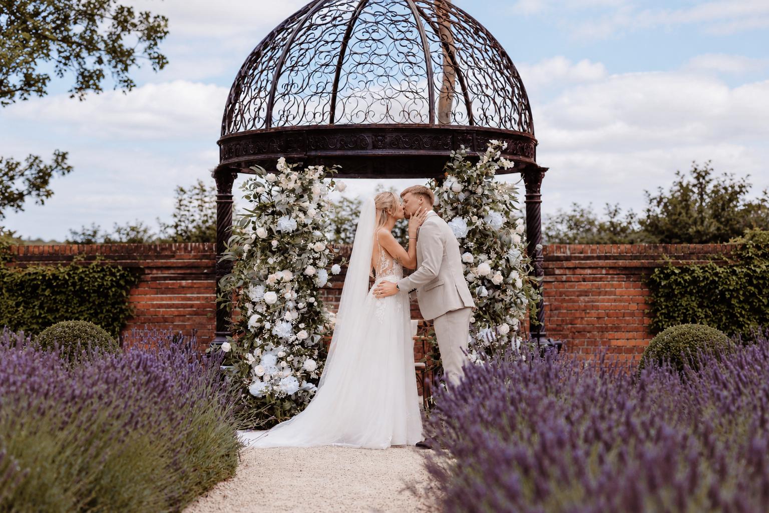 Couple kissing beneath a garden gazebo framed by white and pale blue floral columns, with lavender either side of the aisle