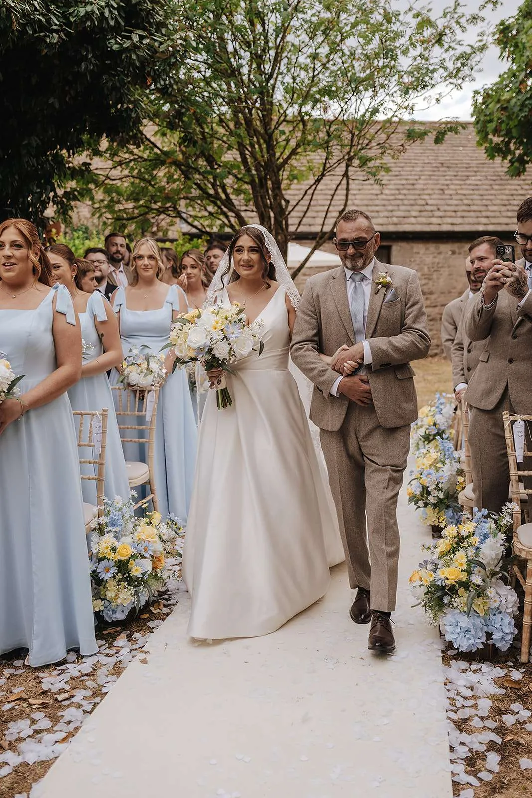 Bride walking down an aisle lined with pale blue and white meadow florals