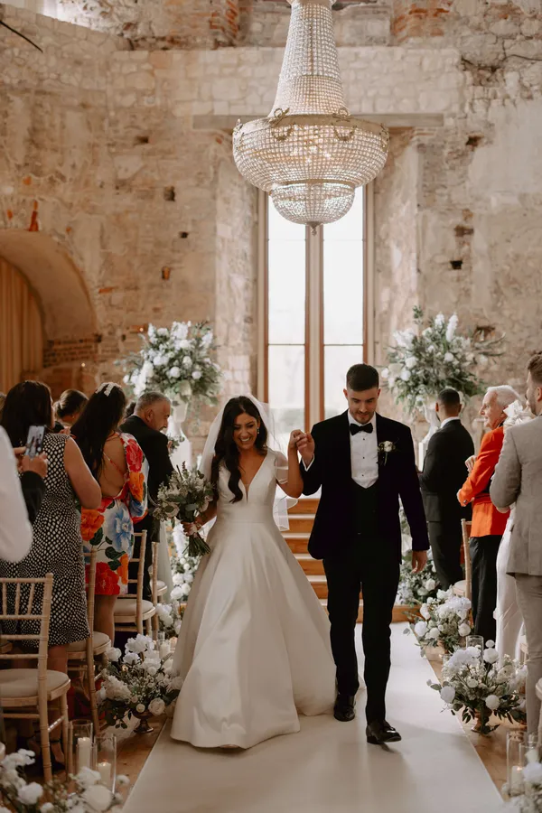 Couple walking back down an aisle at Lulworth Castle lined on both sides with white meadow arrangements
