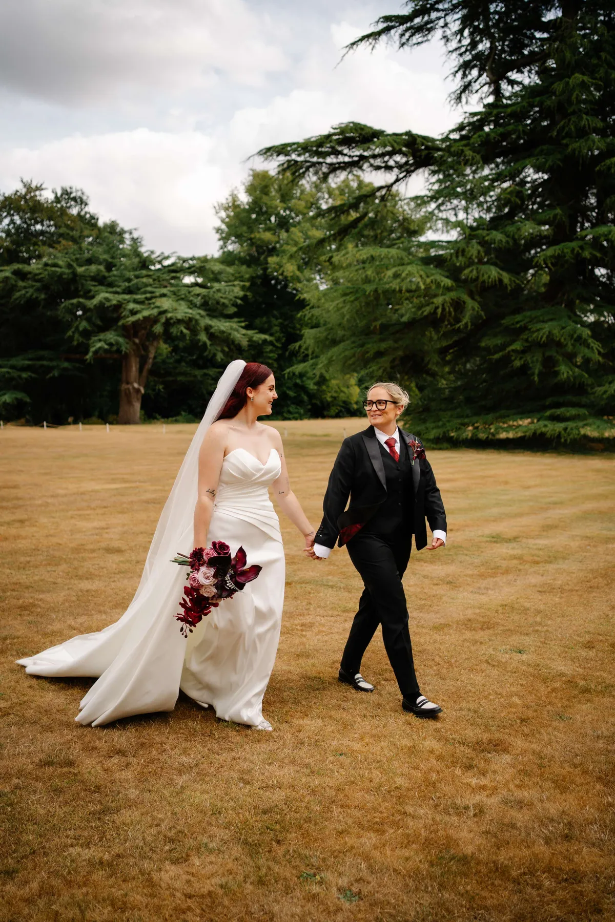Couple walking across a lawn, the bride holding a deep red bouquet