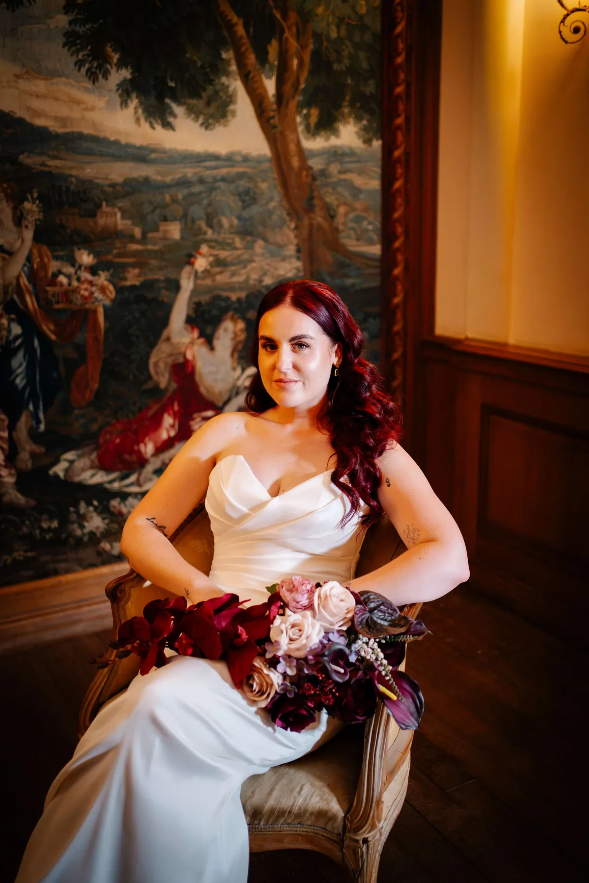 Bride seated in a grand room holding a bouquet of deep red and burgundy blooms