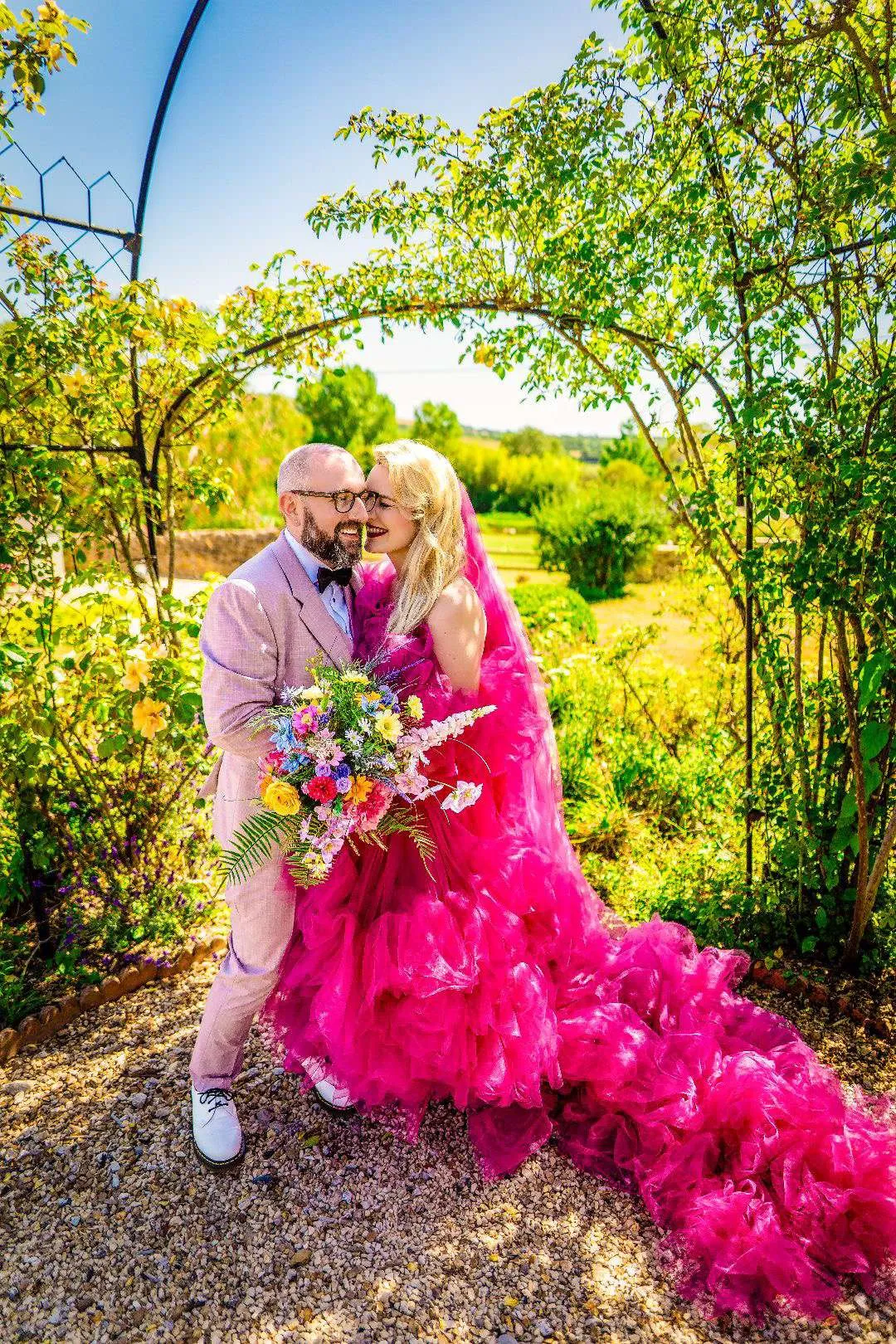 Bride in a bright pink dress with a colourful bouquet, beneath a garden arch