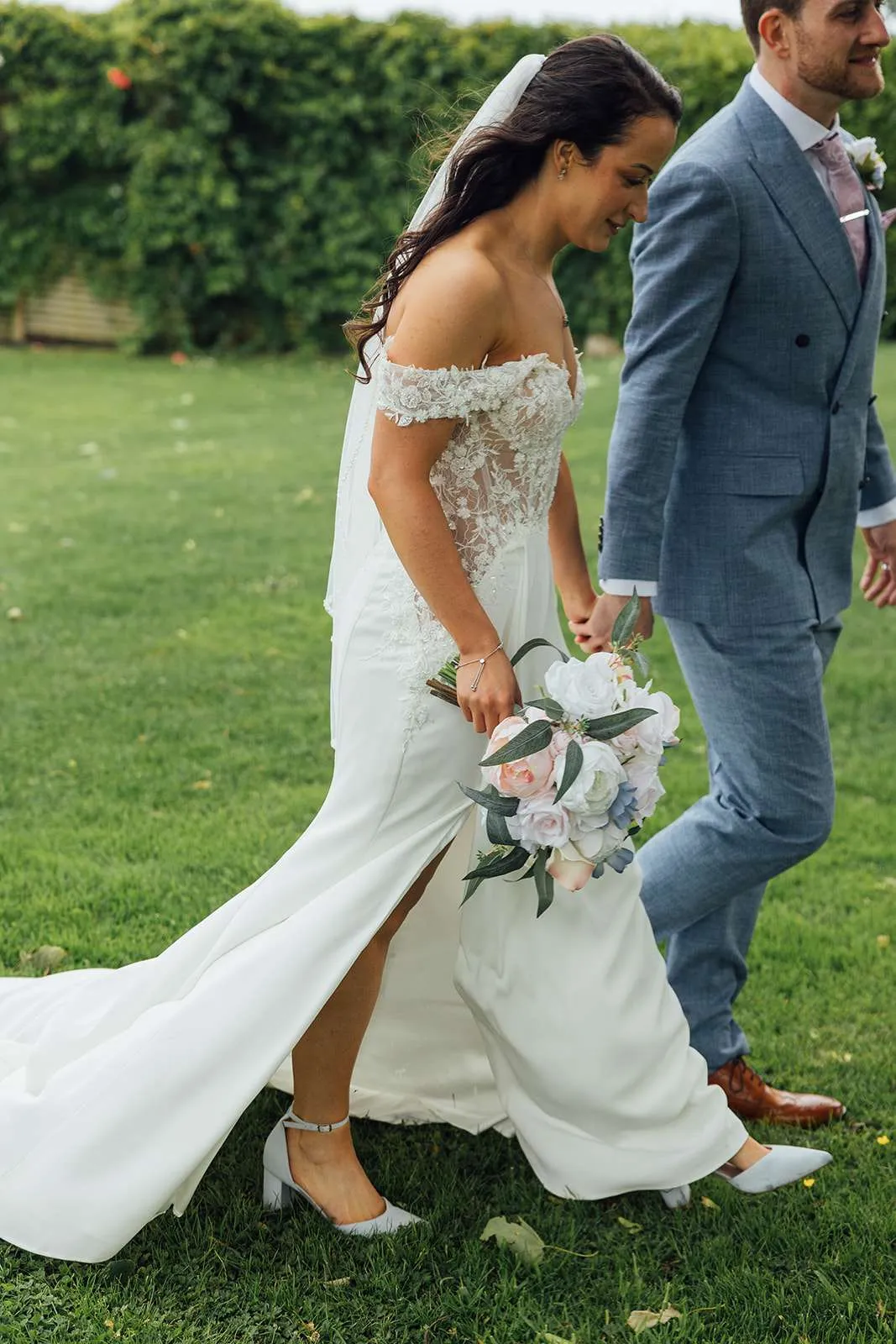 Couple walking across a lawn, the bride holding a pastel bouquet