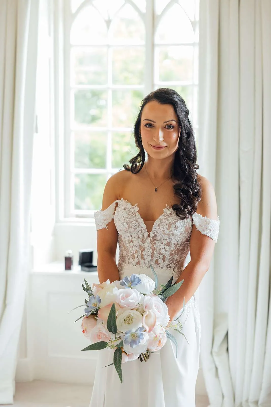 Bride by a tall window holding a soft pastel bouquet of pink, blue and white