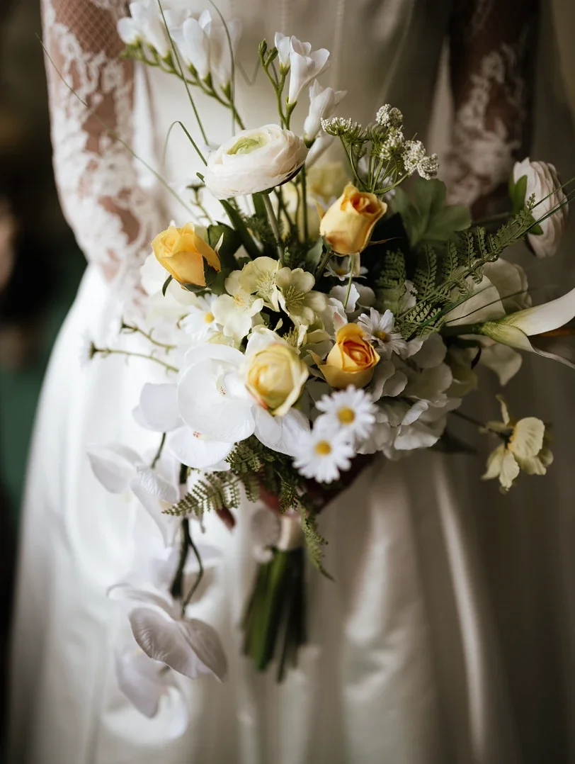Bride in a lace-sleeved dress holding a loose bouquet of white orchids, ranunculus, daisies and soft yellow roses with fern
