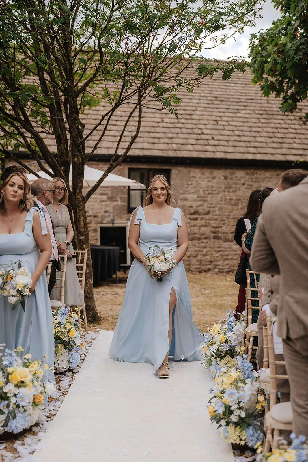 Bridesmaid in pale blue walking down an aisle lined with meadow florals, holding a white and yellow bouquet