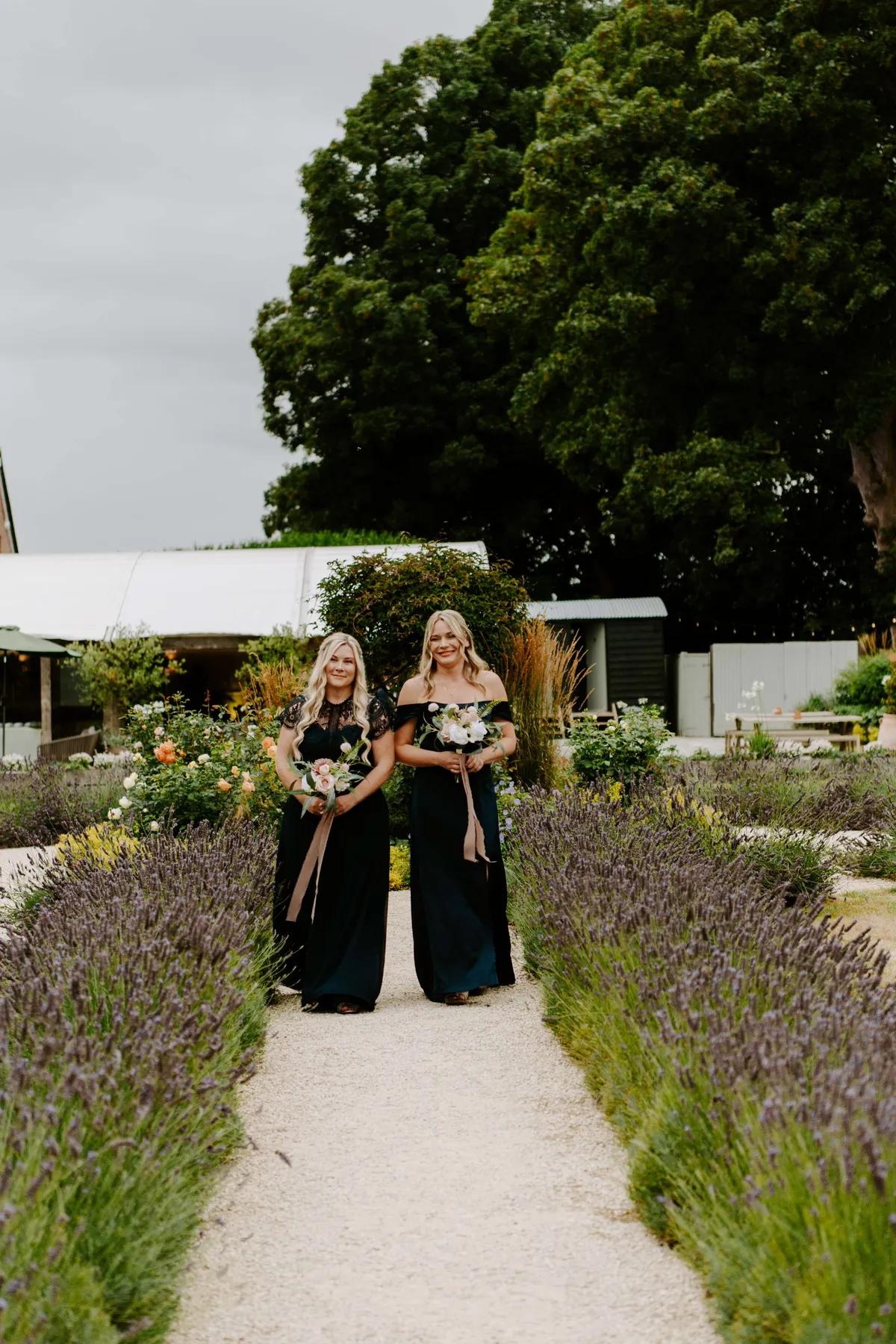 Two bridesmaids in black holding white bouquets on a path lined with lavender