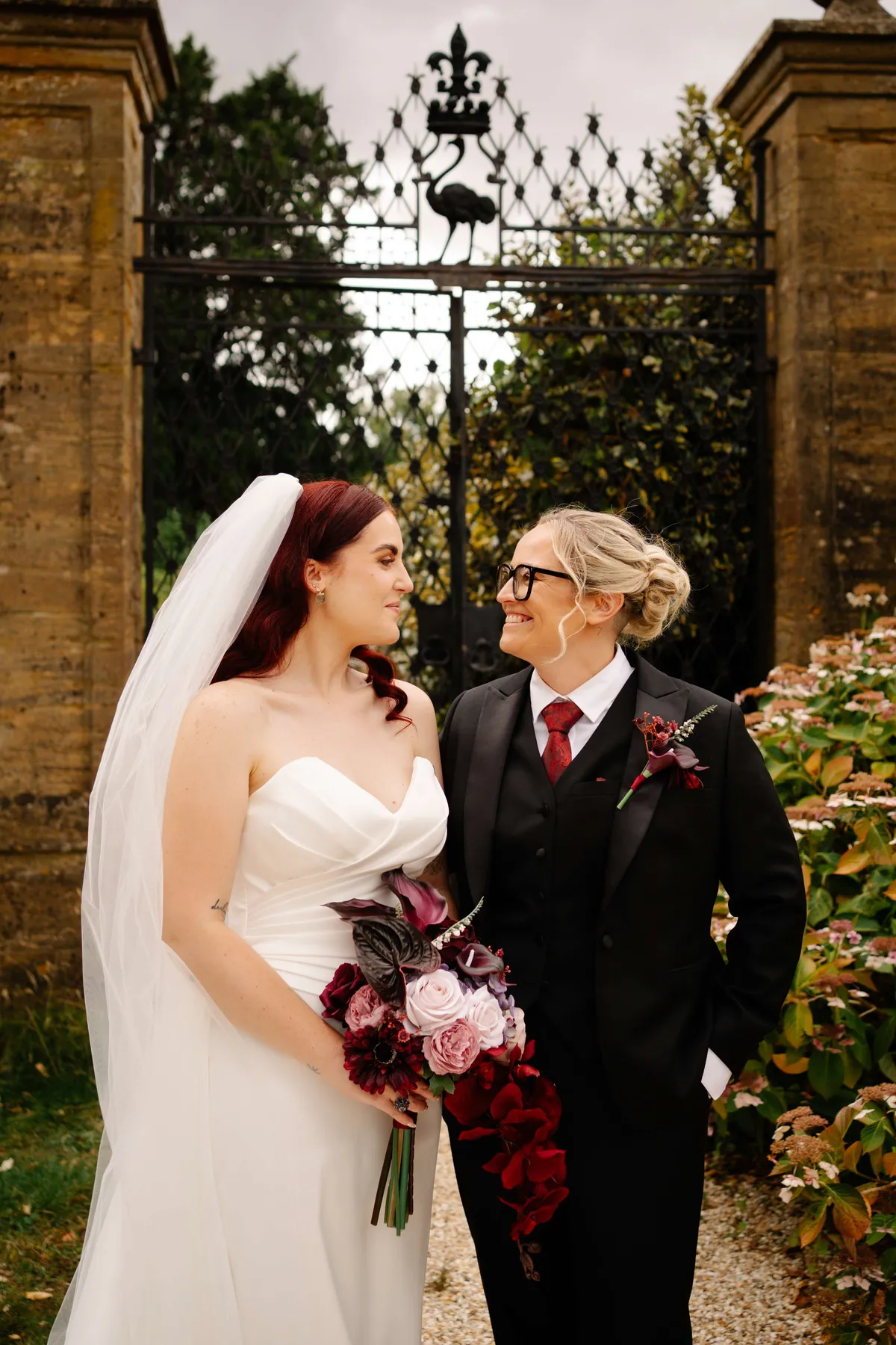 Couple at a wrought iron gate, one wearing a deep red buttonhole to match the bouquet
