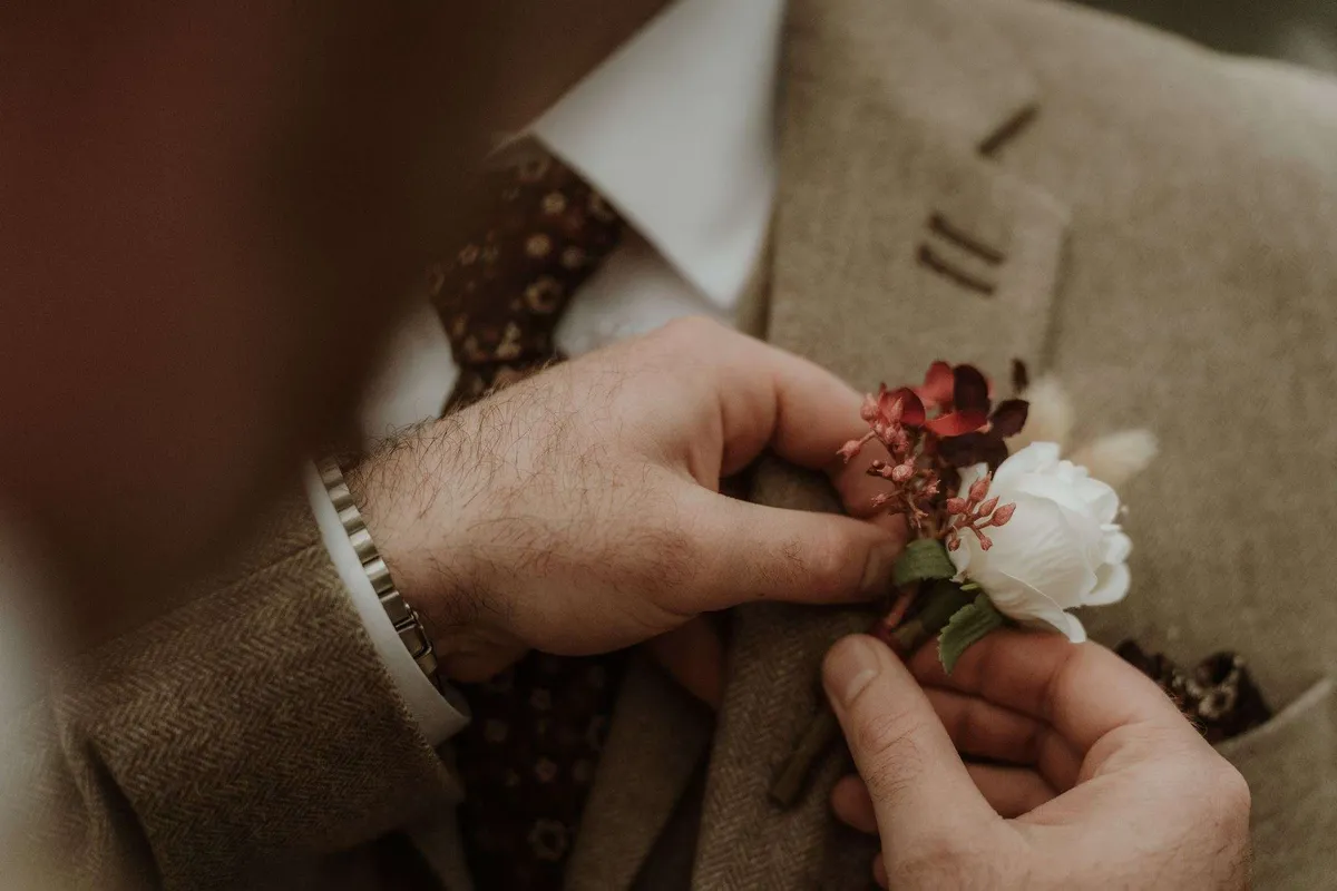 Hands pinning a deep red and white buttonhole onto a tweed jacket