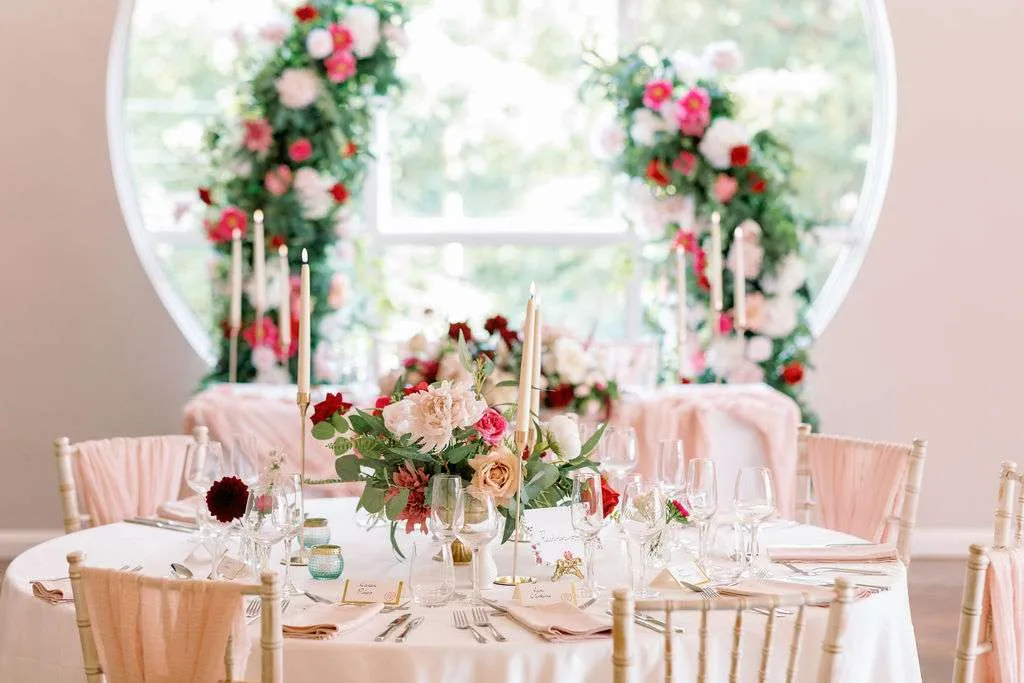 Round table with a pink, red and white footed bowl arrangement and taper candles, floral pillars behind