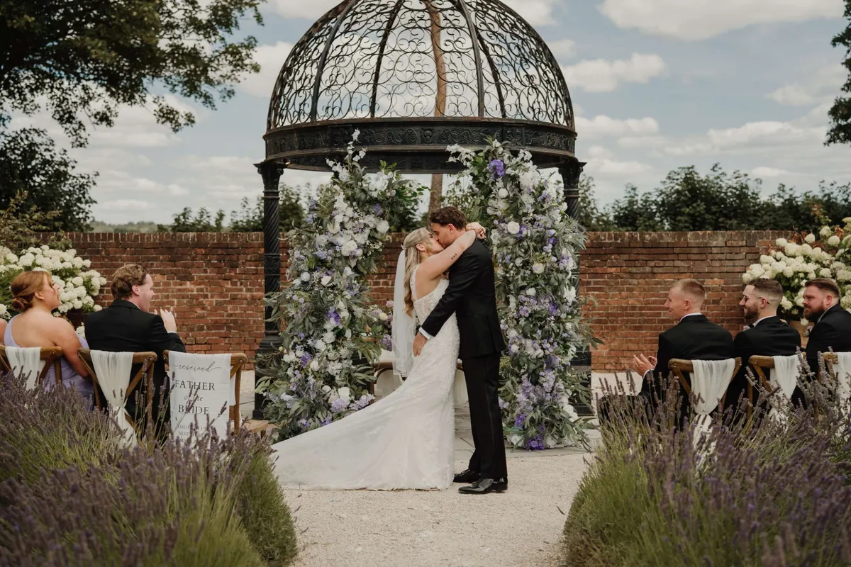 Couple kissing beneath a wrought iron gazebo framed by lilac, white and green floral pillars, with lavender lining the aisle