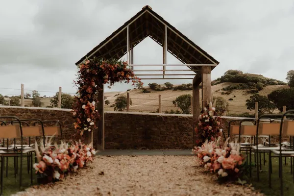 Outdoor ceremony with an oak arbor dressed in rust, burgundy and white florals, the aisle lined with meadow arrangements