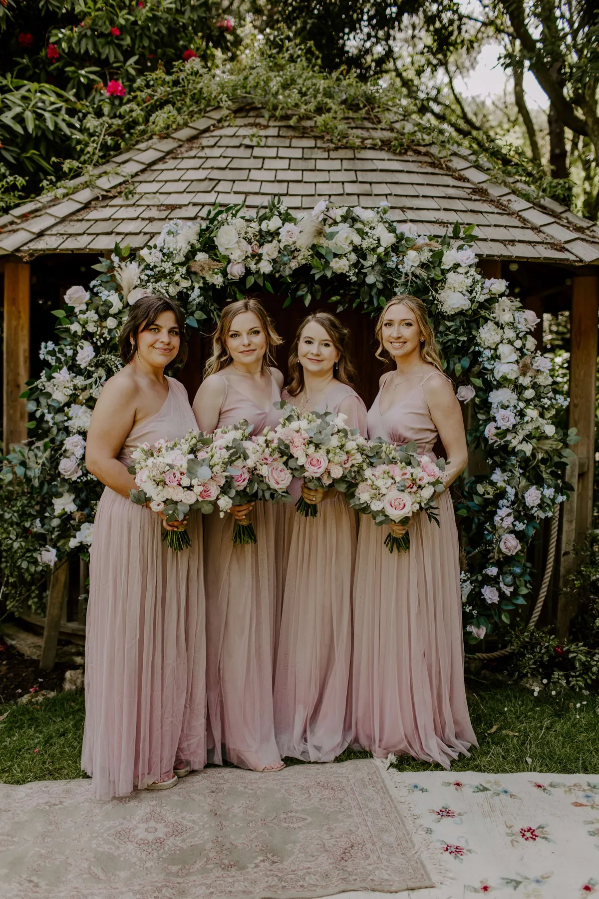 Bridesmaids in blush dresses holding bouquets in front of a floral moongate