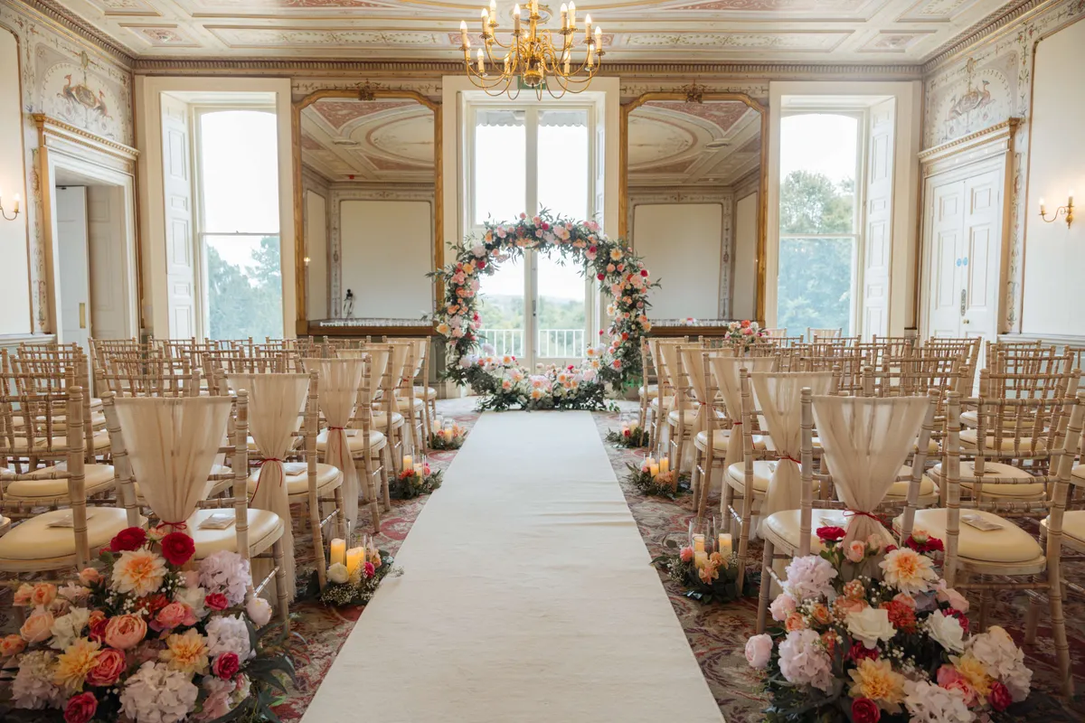 Aisle in a grand hall lined with colourful meadow florals, leading to a floral moongate