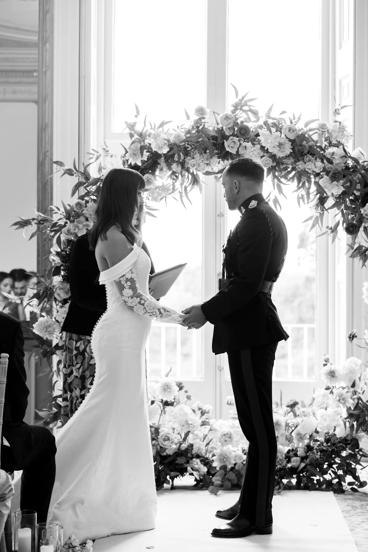 Couple holding hands for their vows in front of a floral moongate, in black and white