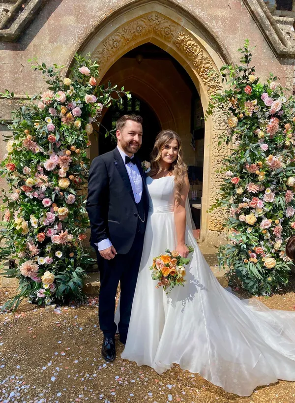Couple outside a church doorway framed by tall pillars of peach, pink and white blooms