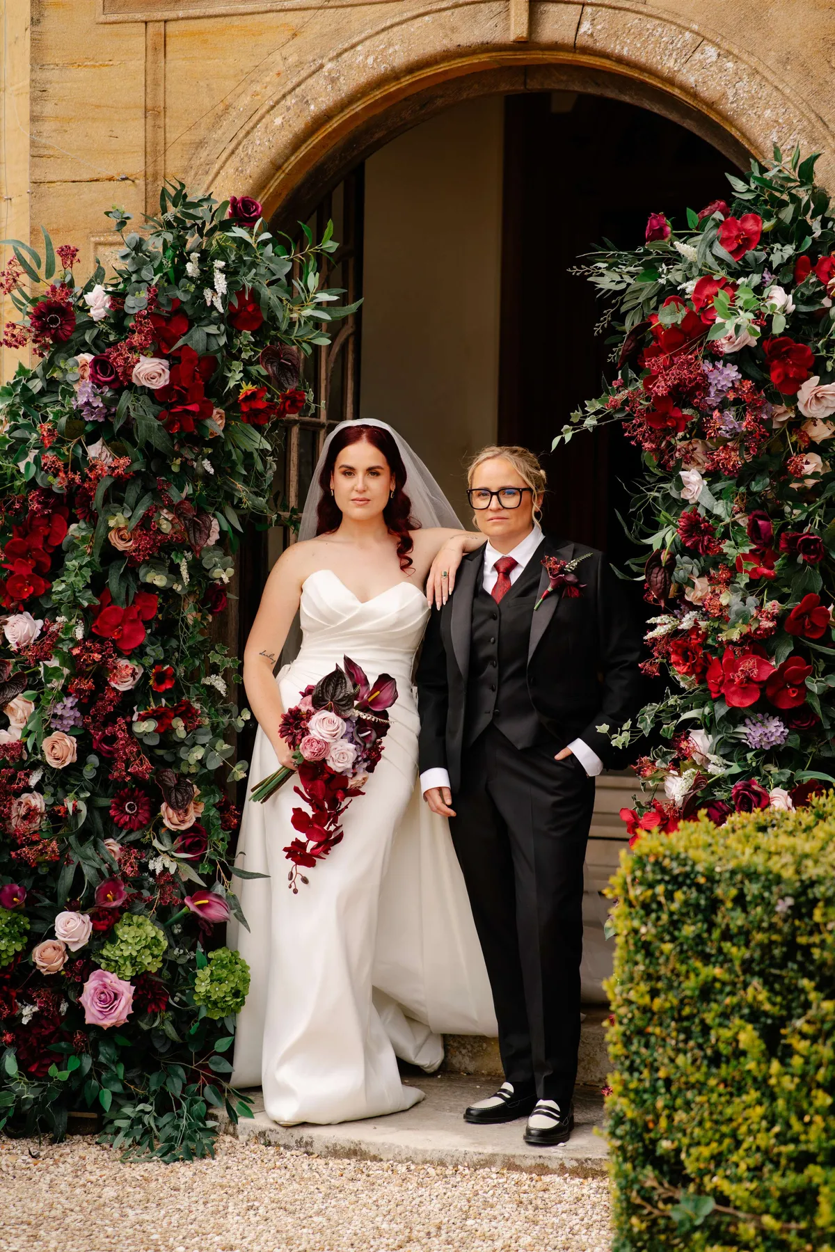 Couple at Minterne House between pillars of deep red, burgundy and pink florals