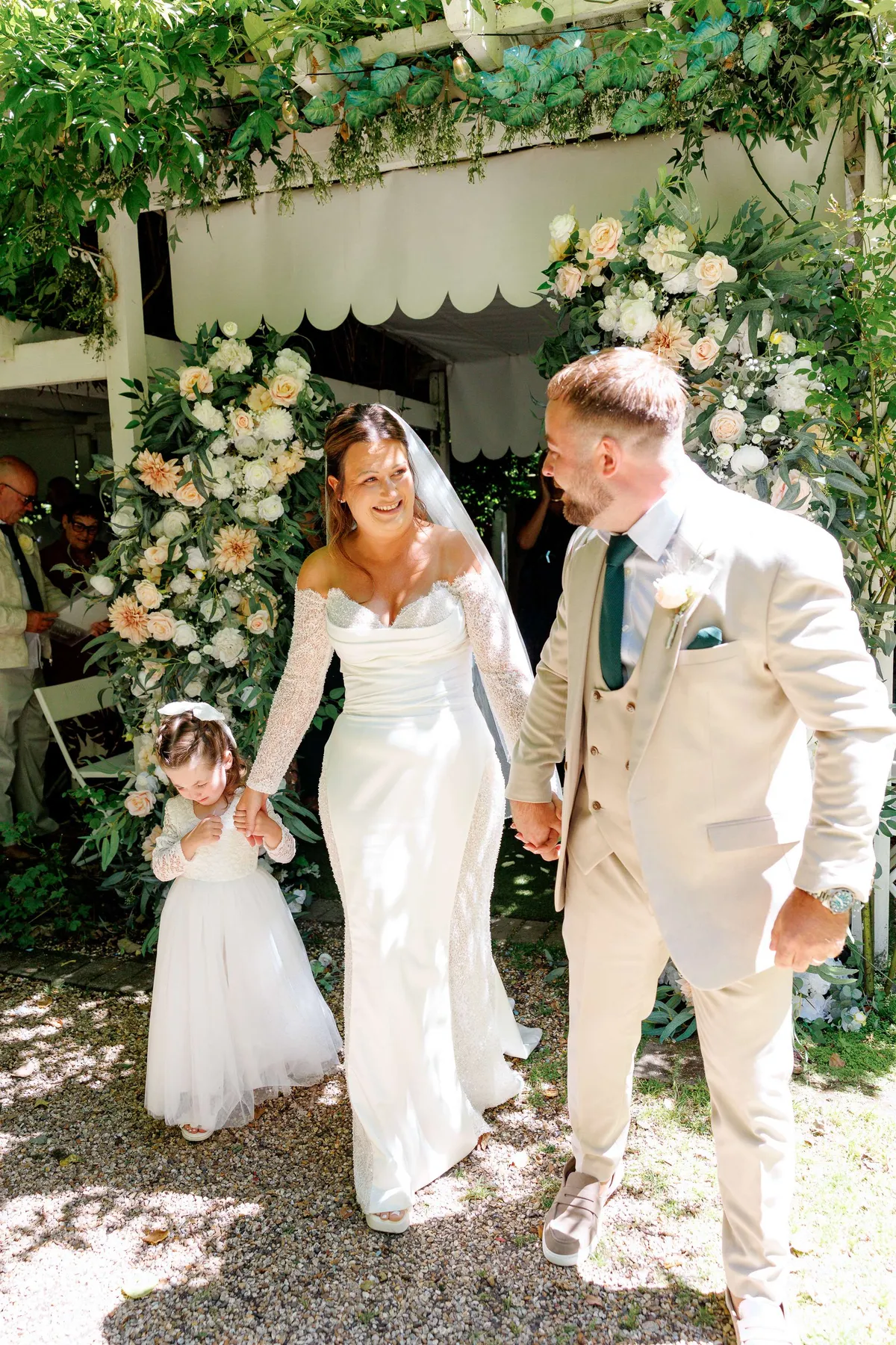 Couple and flower girl walking out between pastel floral pillars at Upwey Wishing Well