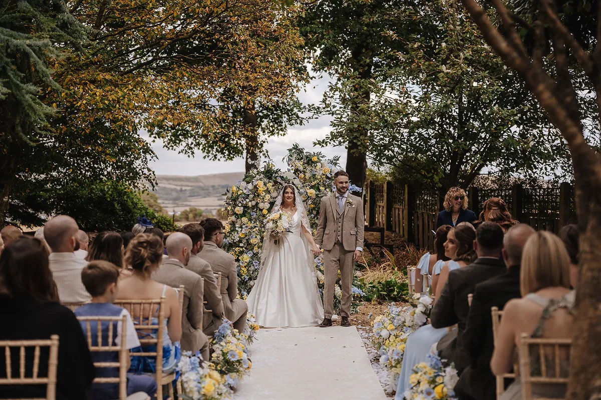 Couple walking back down a garden aisle lined with blue and yellow meadow florals, beneath a floral arch