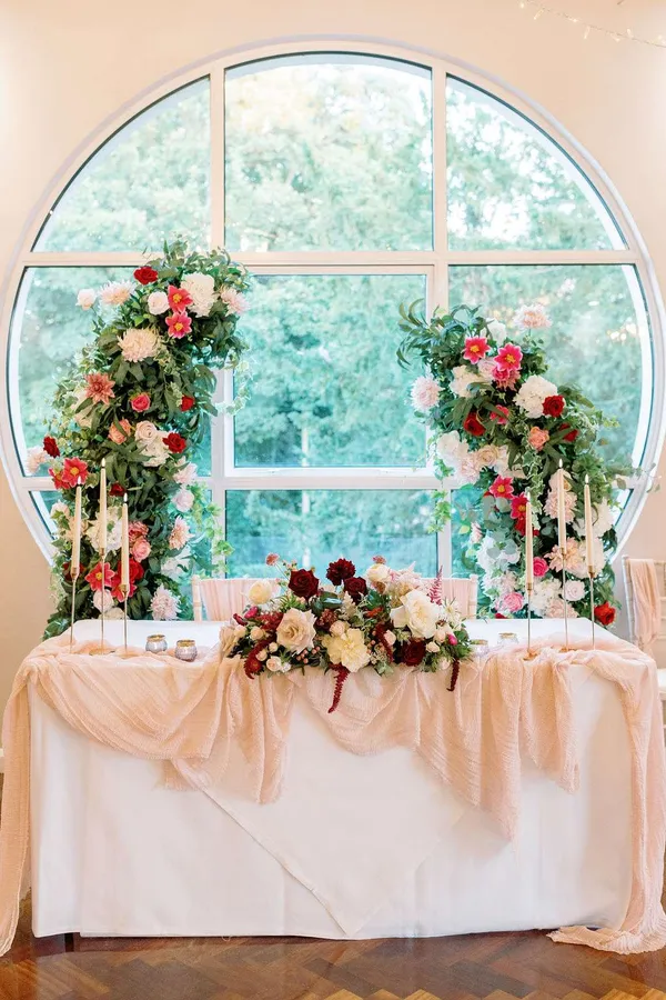 Top table with a pink and red floral arrangement, framed by floral pillars in front of a round window
