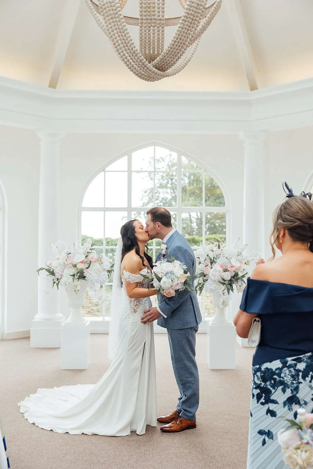 Couple kissing between pastel floral urns on plinths in a bright white ceremony room