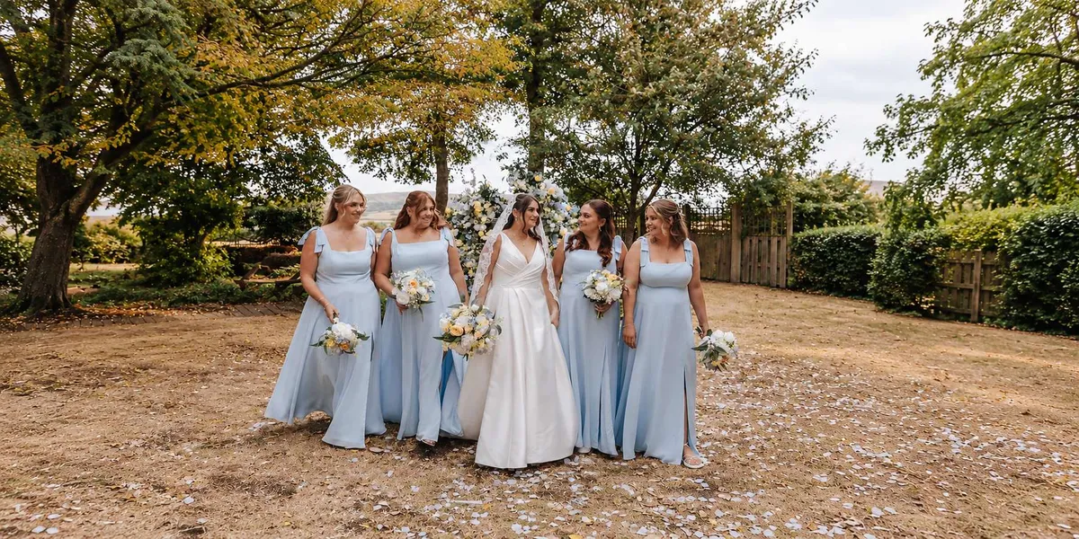 Bride walking with four bridesmaids in pale blue, all holding white, yellow and blue bouquets