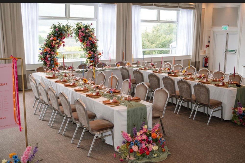 Long reception tables with orange napkins and bright florals beneath a floral arch